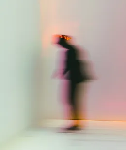 Blurred figure walking indoors against a light-colored wall and floor.