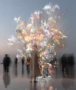 Glowing artificial tree with bright lights surrounded by shadowy figures on a misty surface.